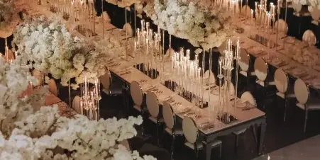 A wedding reception hall featuring long banquet tables with white floral centerpieces and tall, elevated floral installations with taper candles.