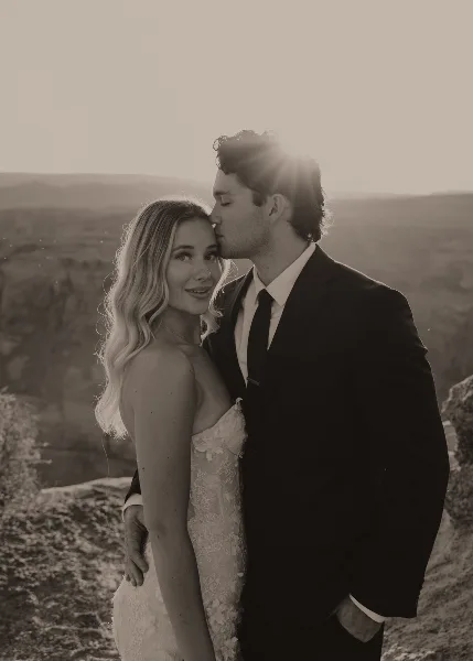 A black and white portrait of a bride and groom embracing outdoors with a scenic landscape background.