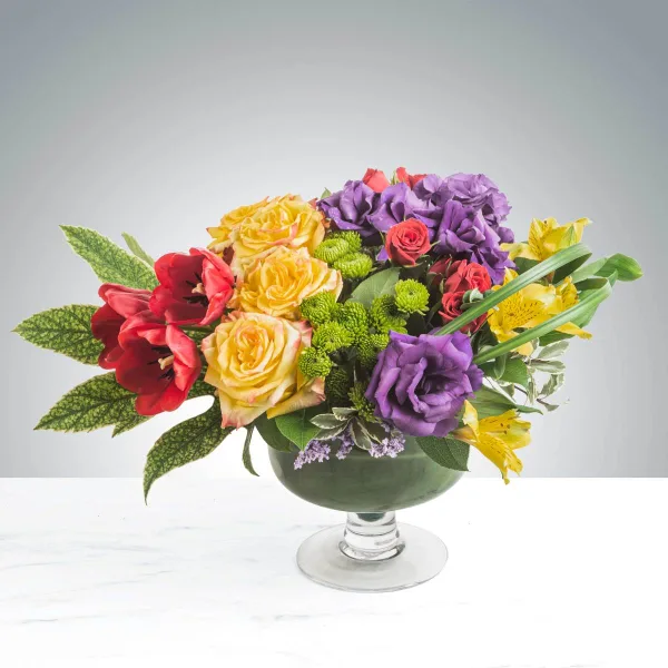 A colorful floral arrangement featuring yellow roses, purple carnations, red amaryllis, and yellow lilies in a green glass pedestal vase.