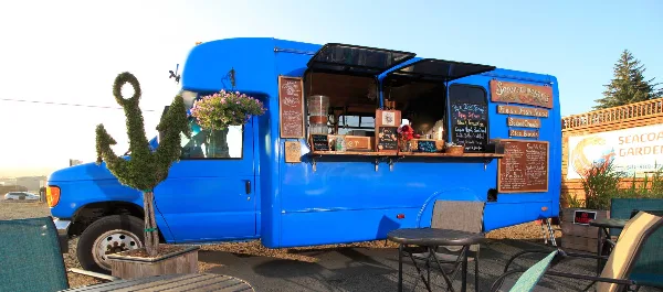 A blue food truck with an open service window, menu boards, and outdoor seating.