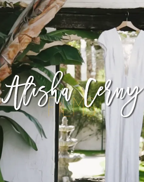 A white wedding dress hanging on a wooden hanger against a backdrop of lush green tropical leaves and a white wall.