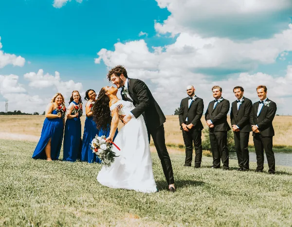 A bride and groom share a dip in a grassy field with their wedding party standing in the background under a blue sky.