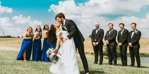 A bride and groom share a dip in a grassy field with their wedding party standing in the background under a blue sky.