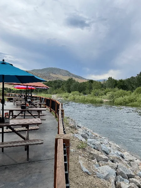 Outdoor wooden picnic tables and umbrellas on a patio overlooking a river with a rocky shoreline and hills in the background.