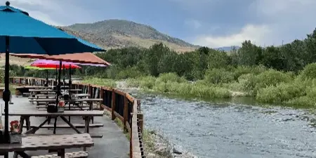 Outdoor wooden picnic tables and umbrellas on a patio overlooking a river with a rocky shoreline and hills in the background.