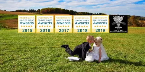 Two children in wedding costumes sitting on a grassy field with award badges overlaid.
