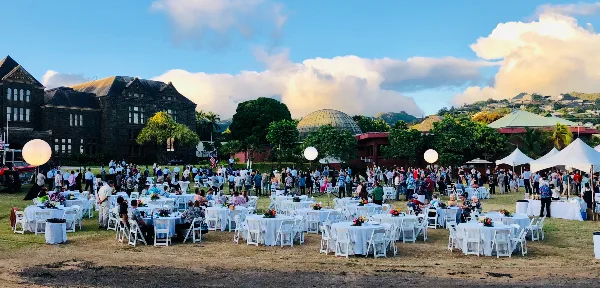 Outdoor event setup with white tables and chairs on a lawn in front of the Bishop Museum in Honolulu.