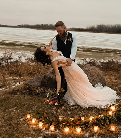 A bride and groom posing in a romantic outdoor setting with a circle of lit candles on the ground.