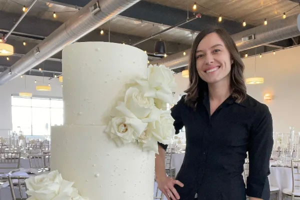 A smiling woman in a black shirt stands next to a tall, white two-tier wedding cake decorated with white sugar roses in a banquet hall.