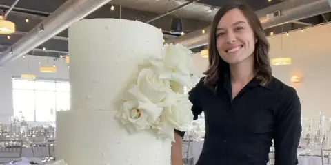 A smiling woman in a black shirt stands next to a tall, white two-tier wedding cake decorated with white sugar roses in a banquet hall.