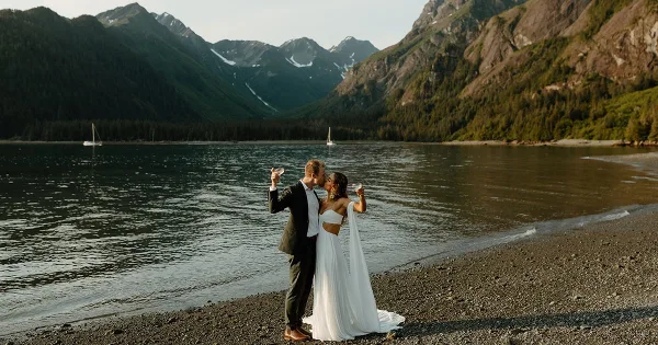 A bride and groom in formal wedding attire standing on a pebble beach by a lake with mountains in the background, holding champagne flutes.