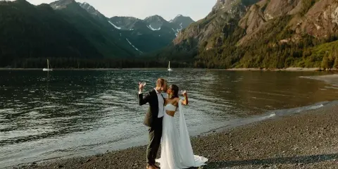 A bride and groom in formal wedding attire standing on a pebble beach by a lake with mountains in the background, holding champagne flutes.