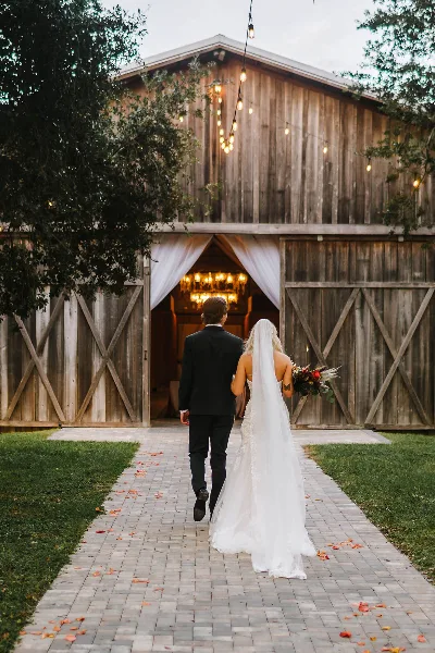 A bride and groom walking down a brick path toward a rustic wooden barn with string lights and white drapery.