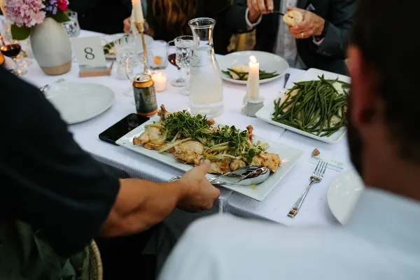 A caterer serving a plated meal of roasted chicken and green beans to guests at a formal event table.
