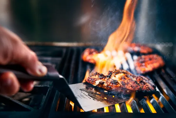 A close-up of a hand using a spatula to flip meat on a hot grill with visible flames.