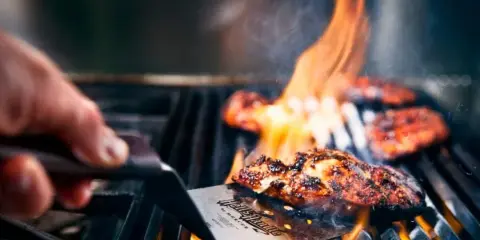 A close-up of a hand using a spatula to flip meat on a hot grill with visible flames.