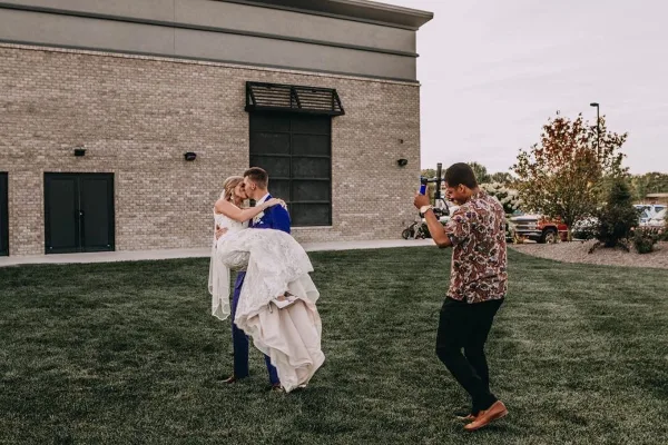 A videographer filming a bride and groom kissing outdoors in front of a brick building.