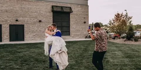 A videographer filming a bride and groom kissing outdoors in front of a brick building.