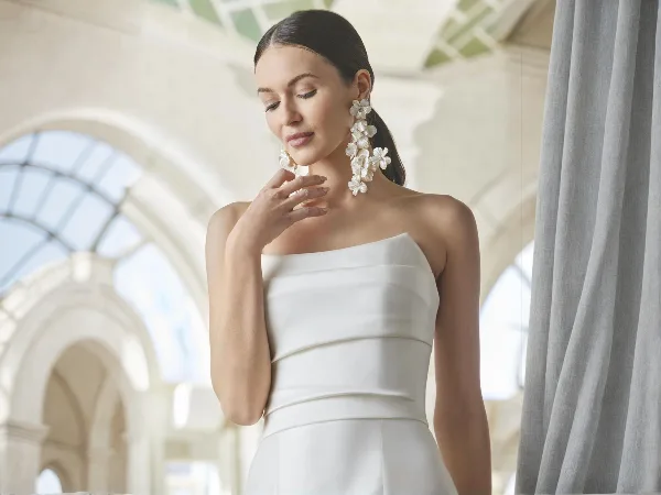 A bride in a white strapless gown with styled hair and makeup, wearing floral earrings, standing in an arched interior.