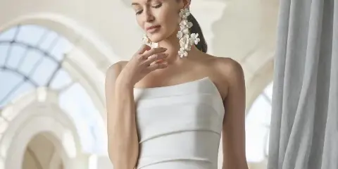 A bride in a white strapless gown with styled hair and makeup, wearing floral earrings, standing in an arched interior.