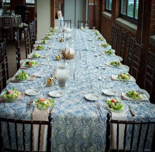A long banquet table set with blue patterned tablecloth, white plates, salad bowls, and glassware in a brick-walled venue.