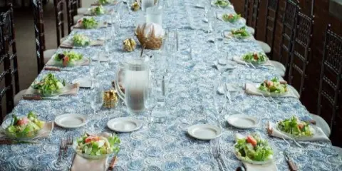 A long banquet table set with blue patterned tablecloth, white plates, salad bowls, and glassware in a brick-walled venue.