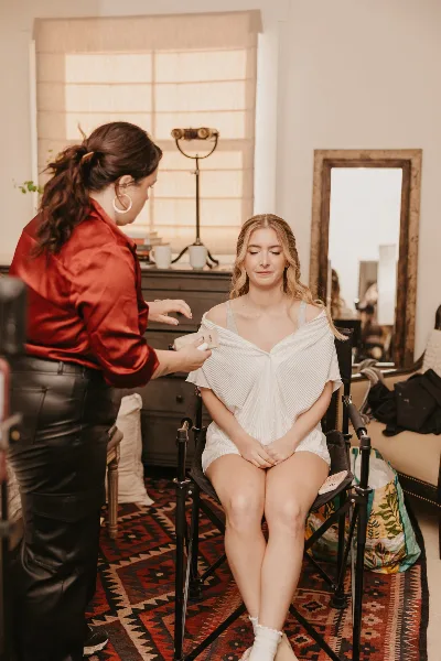 A makeup artist in a red shirt applying makeup to a seated bride in a white dress.
