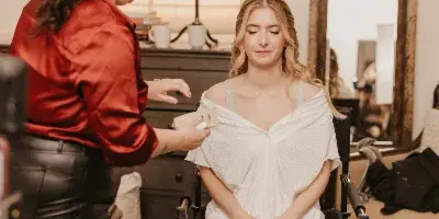 A makeup artist in a red shirt applying makeup to a seated bride in a white dress.