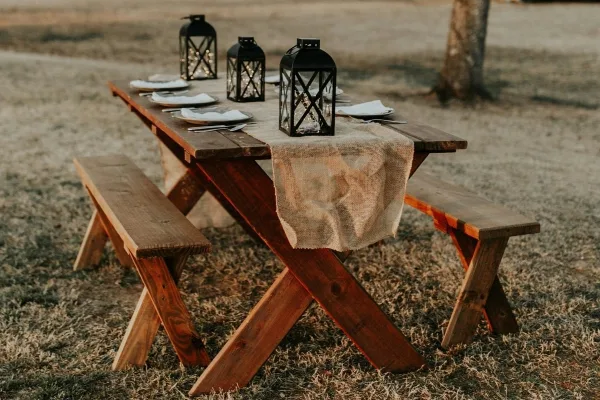 Rustic wooden table and benches set outdoors with lanterns and place settings.