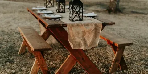 Rustic wooden table and benches set outdoors with lanterns and place settings.
