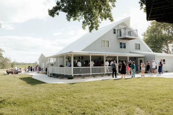 A white barn-style wedding venue with a covered porch where a group of people are gathered on a grassy lawn.