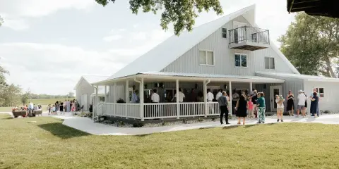 A white barn-style wedding venue with a covered porch where a group of people are gathered on a grassy lawn.