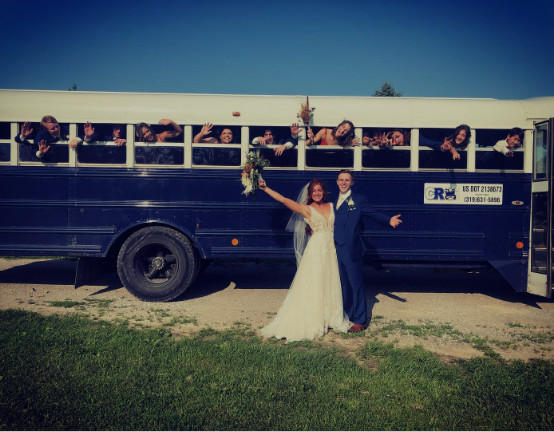 A bride and groom posing in front of a blue party bus with a group of people waving from the windows.