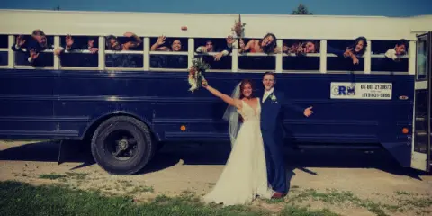 A bride and groom posing in front of a blue party bus with a group of people waving from the windows.