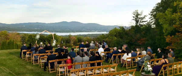 Outdoor wedding ceremony with guests seated on a grassy hill overlooking a lake and mountains.