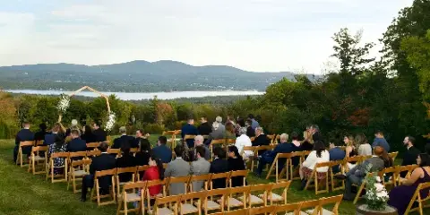 Outdoor wedding ceremony with guests seated on a grassy hill overlooking a lake and mountains.