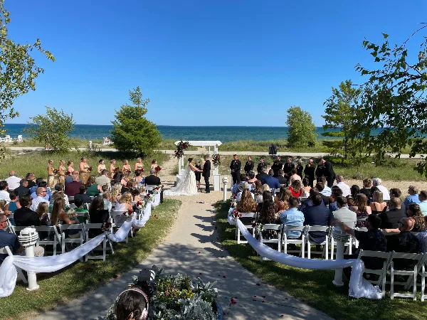 Outdoor wedding ceremony in progress with a bride and groom at an altar, officiant, and seated guests near a body of water.