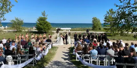 Outdoor wedding ceremony in progress with a bride and groom at an altar, officiant, and seated guests near a body of water.