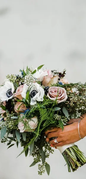A hand holding a lush bridal bouquet with pink roses, white anemones, and greenery.