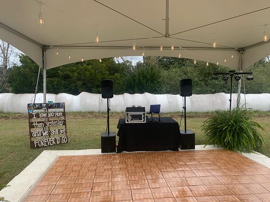A wedding event setup under a white tent featuring a wooden dance floor, professional speakers, and a sign reading 'FOREVER TO GO'.