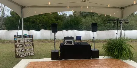 A wedding event setup under a white tent featuring a wooden dance floor, professional speakers, and a sign reading 'FOREVER TO GO'.