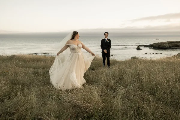 A bride in a white gown and a groom in a dark suit standing in tall coastal grass with the ocean in the background.