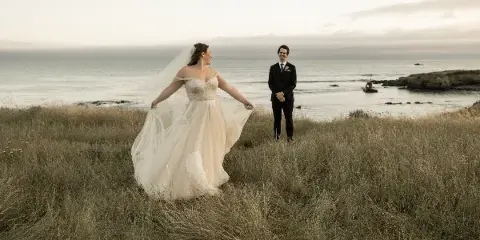 A bride in a white gown and a groom in a dark suit standing in tall coastal grass with the ocean in the background.