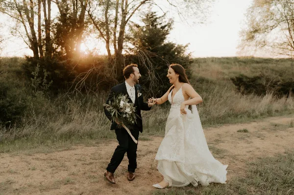 A bride and groom laughing and holding hands in a grassy field with trees and a sunset in the background.