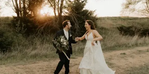 A bride and groom laughing and holding hands in a grassy field with trees and a sunset in the background.