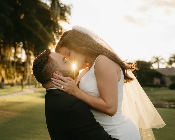 A bride and groom sharing a kiss outdoors at sunset, with the bride wearing a white dress and veil.