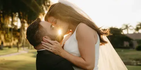 A bride and groom sharing a kiss outdoors at sunset, with the bride wearing a white dress and veil.