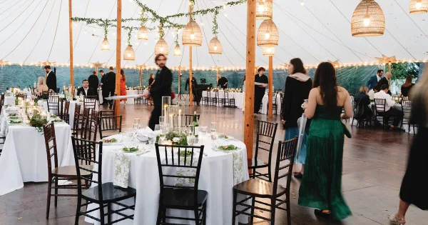 Interior of a large white event tent set up for a wedding reception with round tables, white linens, black chairs, and hanging pendant lights.
