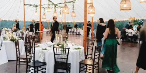 Interior of a large white event tent set up for a wedding reception with round tables, white linens, black chairs, and hanging pendant lights.