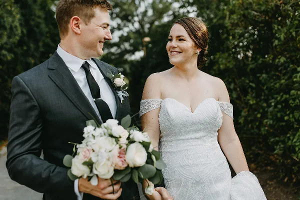 A bride in a white lace gown and a groom in a dark suit holding a bouquet of white and pink flowers, smiling at each other outdoors.